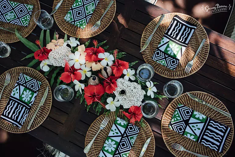 A table setting with six woven round placemats, each with a black, white, and green patterned napkin, silverware, and water glasses. The center of the table has a floral arrangement with red and white flowers, green leaves, and white decorative candl