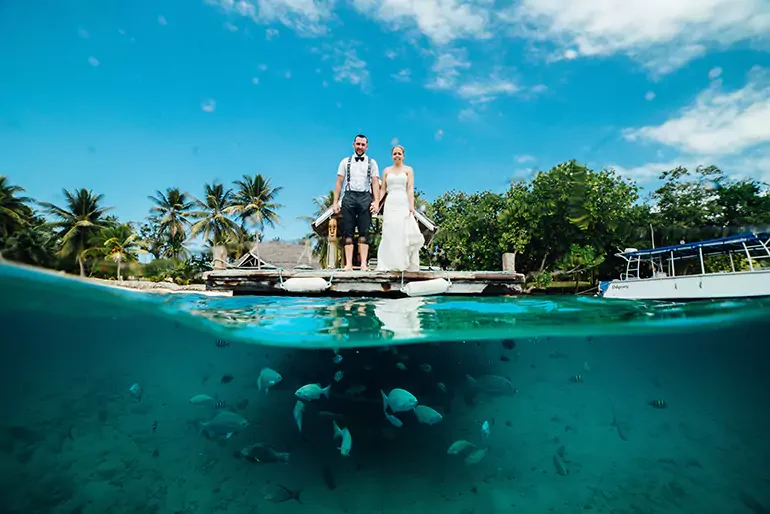 A man and woman in wedding attire standing on a dock by the ocean, with palm trees and a boat nearby, under a partly cloudy blue sky. The photo captures both above and below water views, showing fish swimming underwater.