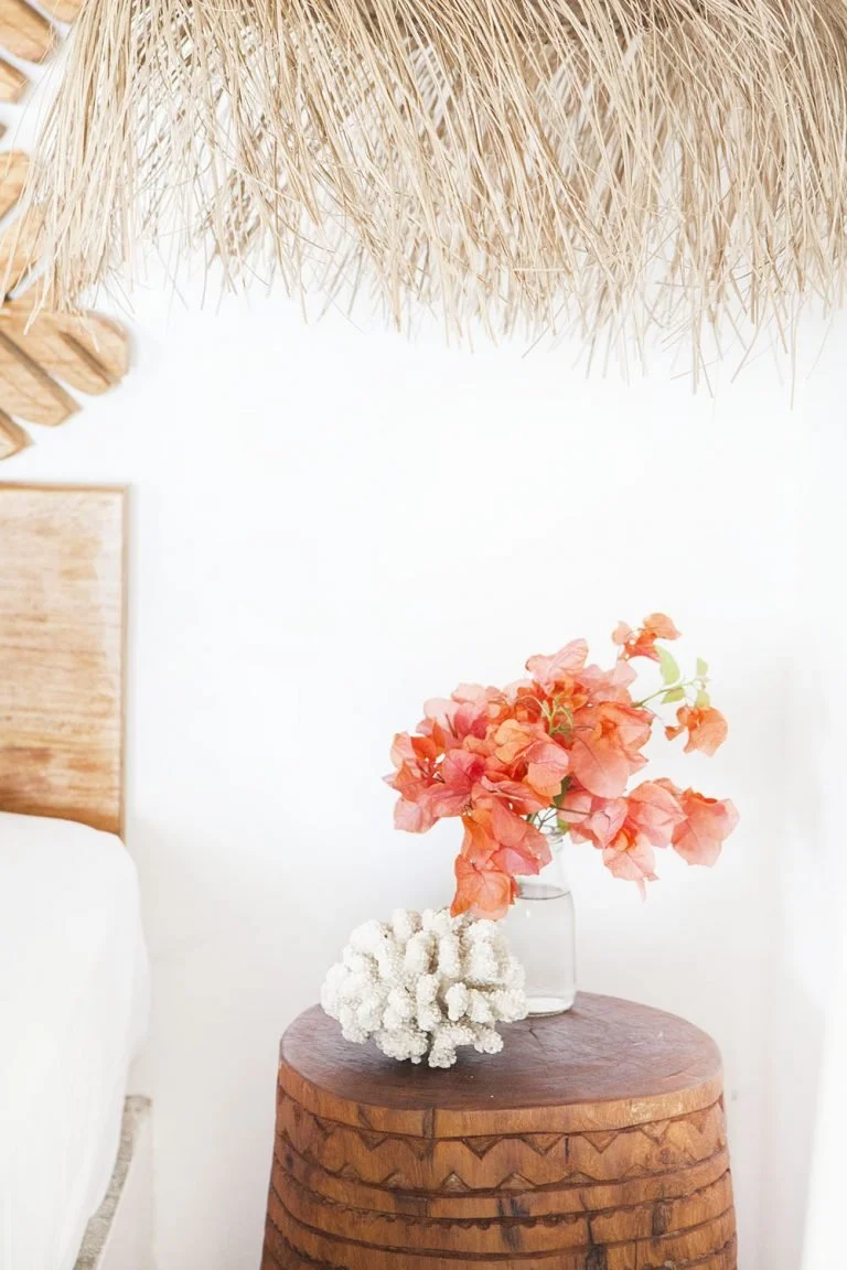 A wooden side table with a pinkish-orange flower arrangement in a clear vase and a white coral sculpture on top, against a white wall with a woven hanging above.