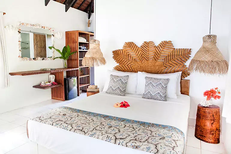 A tropical bedroom with white walls and a wooden leaf-shaped headboard. There is a white bed with two pillows and flower petals on top. Two hanging woven lamps flank the bed. To the right, a round wooden side table with a small bouquet of red flowers