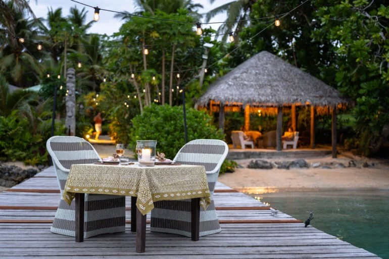 Outdoor dining setup with a round table and two chairs on a wooden deck, with a tropical hut, lush greenery, and string lights in the background.