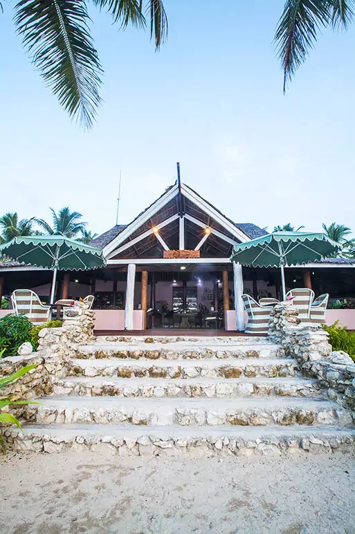 A tropical beachside restaurant with stone steps leading up to an open pavilion, surrounded by palm trees and shaded seating areas.