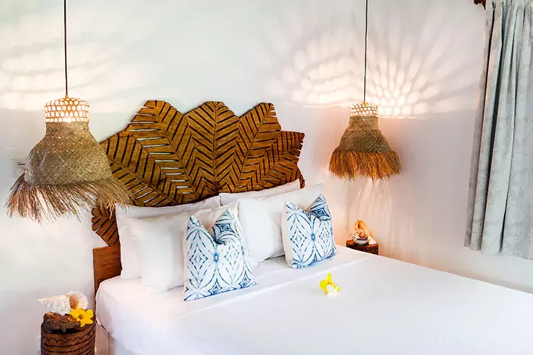 A neatly made bed with white linens and decorative blue pillows, a wooden headboard with leaf-shaped design, two hanging wicker pendant lights, and small bedside tables with yellow flower decorations in a bright, tropical-styled room.