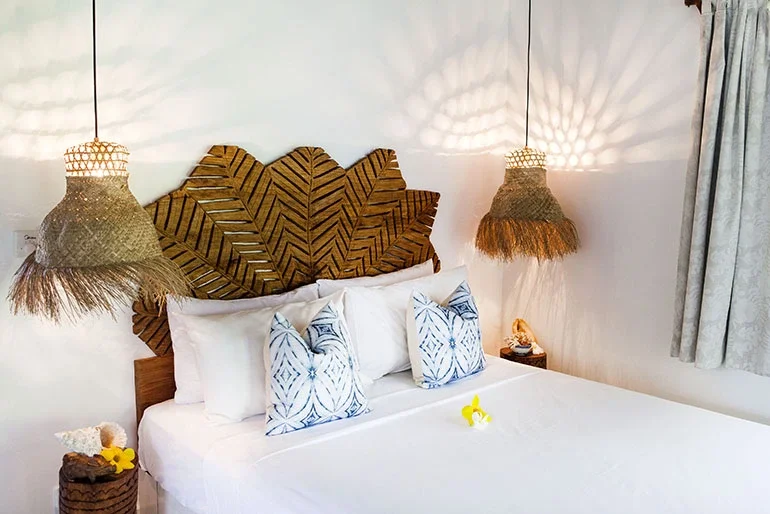 A neatly made bed with white bedding and three white pillows with blue patterns, a tropical leaf-themed headboard, hanging woven pendant lights, a nightstand with a lamp and decorative items, and a window with light-colored curtains.
