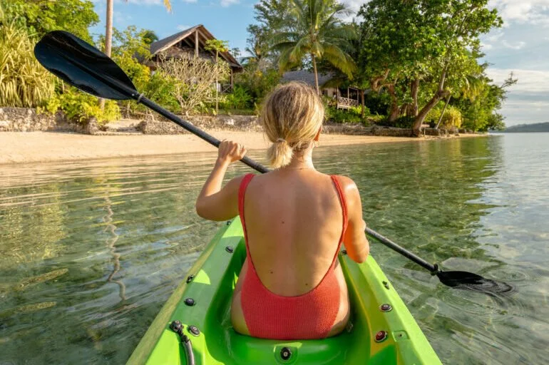A woman with blonde hair tied in a ponytail paddles a green kayak on calm water near a sandy shore with lush trees and a wooden house in the background.