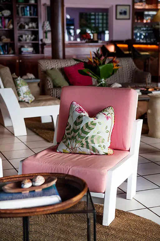 A cozy, pink upholstered chair with a decorative pillow featuring a green leafy pattern in a warm, inviting living room with wooden accents, a glass-top table, and a bouquet of flowers in the background.