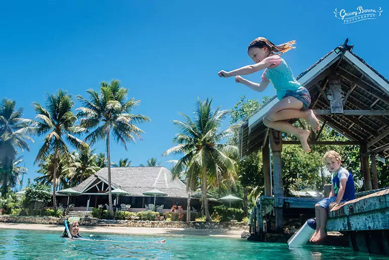 A young girl in a blue shirt jumps off a wooden dock into the water, while a boy sits on the dock, and an older woman floats in the water nearby in a tropical setting with palm trees and a hut.