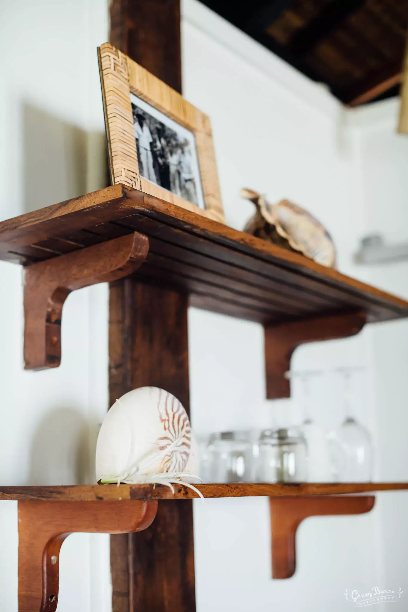 A wooden wall-mounted shelf holds a fish-shaped ceramic piggy bank and a black-and-white framed photograph. The shelf has a rustic finish with visible wood grain and curved support brackets. The room has white walls, and other shelves and glassware a