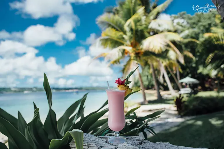 A pink cocktail with a pineapple slice and a flower garnish on a tropical island with palm trees and a beach in the background.
