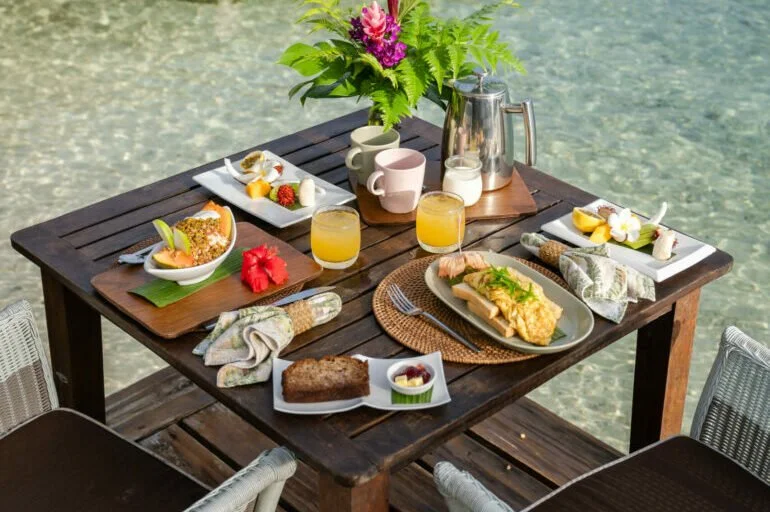 Breakfast table set outdoors by the water with plates of food, glasses of orange juice, a teapot, coffee mugs, and utensils, decorated with a flower arrangement.
