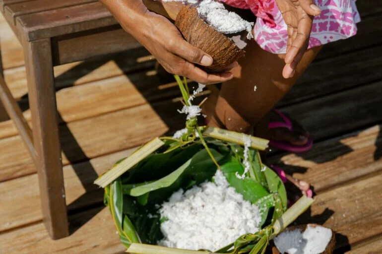 Child using a wooden spoon to sprinkle shredded coconut from a coconut shell into a banana leaf bowl filled with shredded coconut, on a wooden deck.