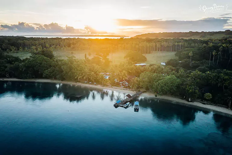 A tropical coastal scene at sunset with a sandy beach, lush green trees, and a small dock with boats on calm water.