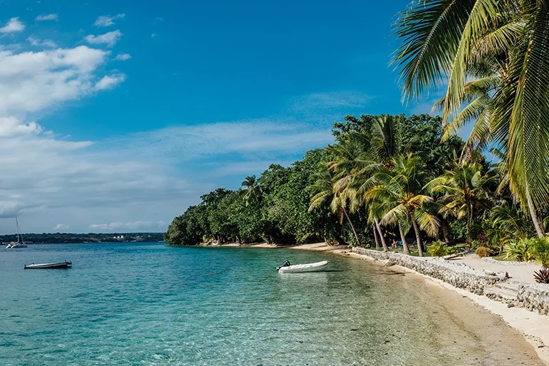 Tropical beach with palm trees, clear blue water, boats, and a partly cloudy sky.