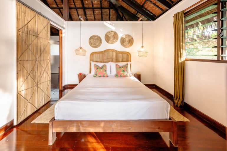 A cozy bedroom with a wooden bed frame, white bedding, and colorful throw pillows. The room has wooden floors, a large window with yellow curtains, and decorative wall hangings above the bed. There are two hanging pendant lights on either side of the