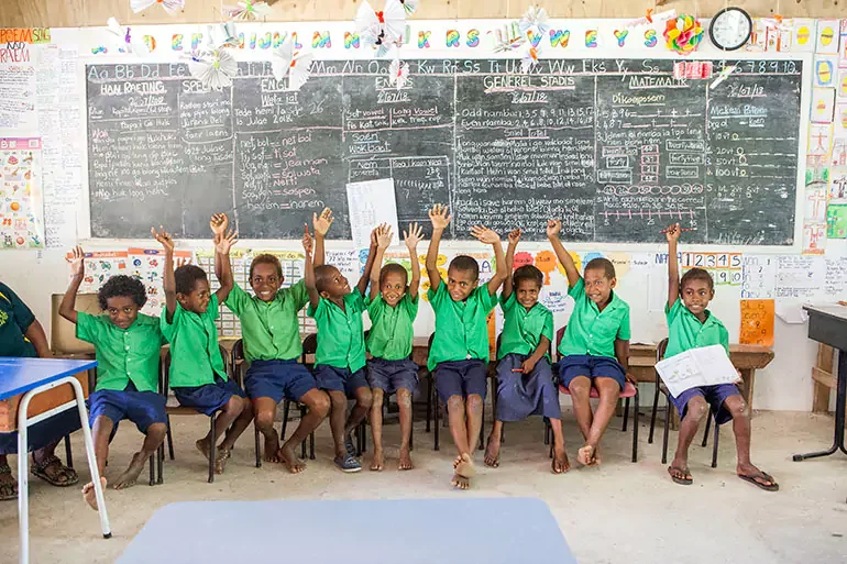 A group of young students in green shirts and dark shorts sitting on chairs in a classroom, raising their hands. The background features a large chalkboard filled with writing and drawings, colorful decorations, and learning posters.