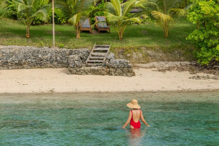 A woman in a red swimsuit and straw hat wades in clear water near a sandy beach, with green palm trees and seating area with chairs on a grassy hill in the background.
