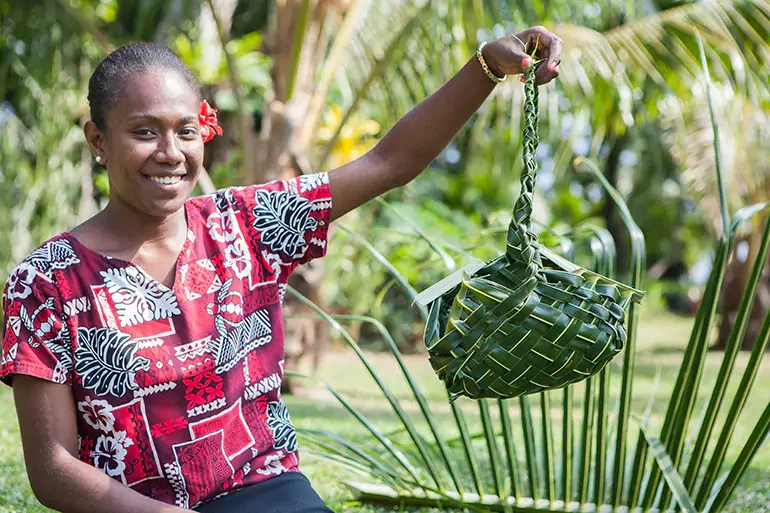 A young woman in a red and white patterned dress holds up a woven basket made from palm leaves outdoors, smiling at the camera, with lush green plants in the background.