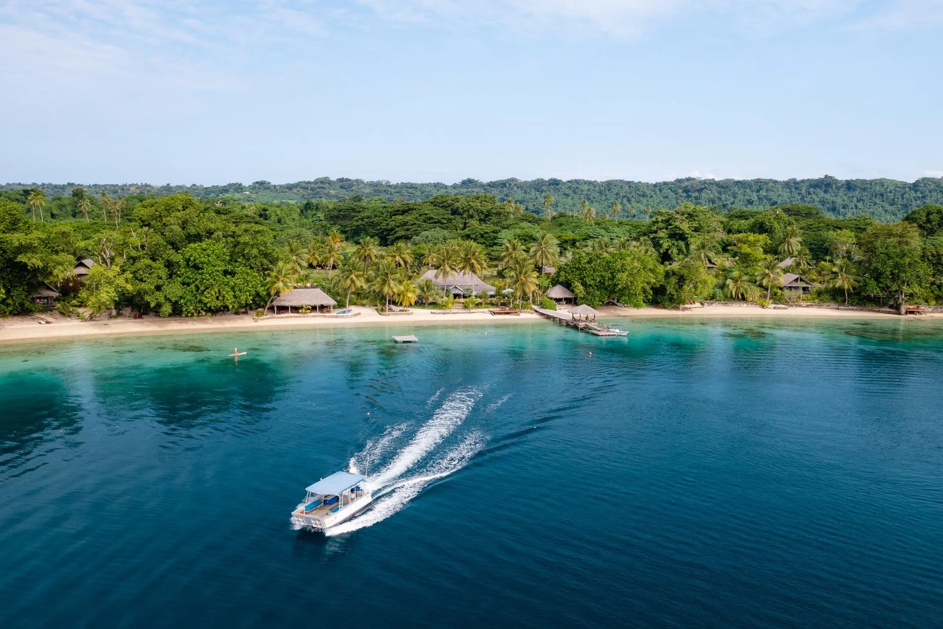 A boat speeds through clear blue water near a tropical beach with palm trees and thatched-roof huts, lush green forest in the background, and a partly cloudy sky above.