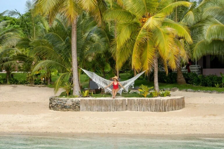 A woman sitting on a hammock on a tropical beach, surrounded by palm trees and sandy shore.