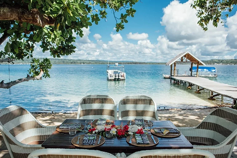 A waterfront dining setup featuring a table with plates and utensils, surrounded by chairs, overlooking a lake with a boat and a small dock under a partly cloudy sky.