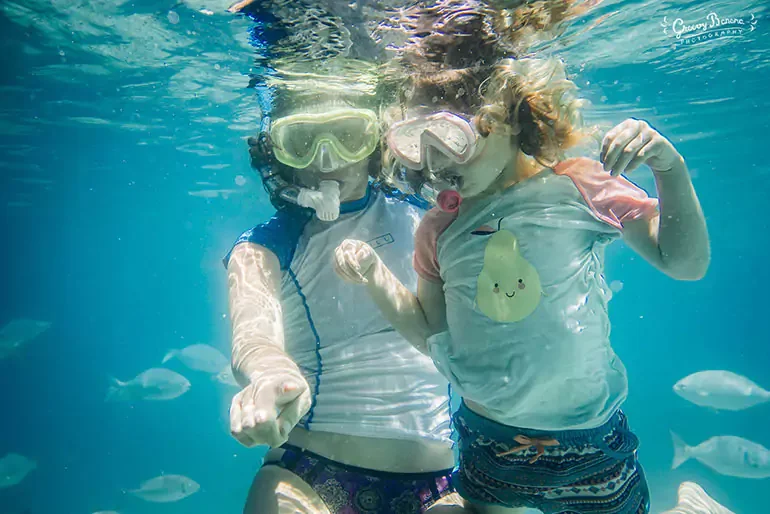 Two children wearing snorkeling gear and swimming underwater, surrounded by fish.