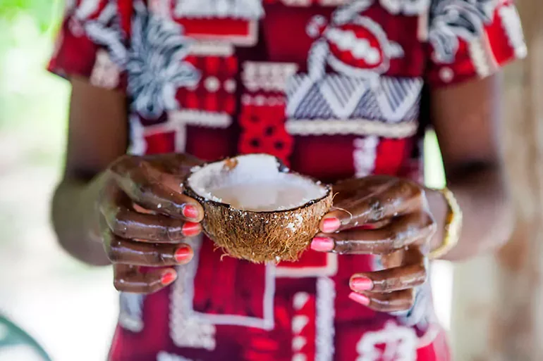 Person holding a split open coconut with white flesh inside, wearing a colorful patterned shirt and painted fingernails.