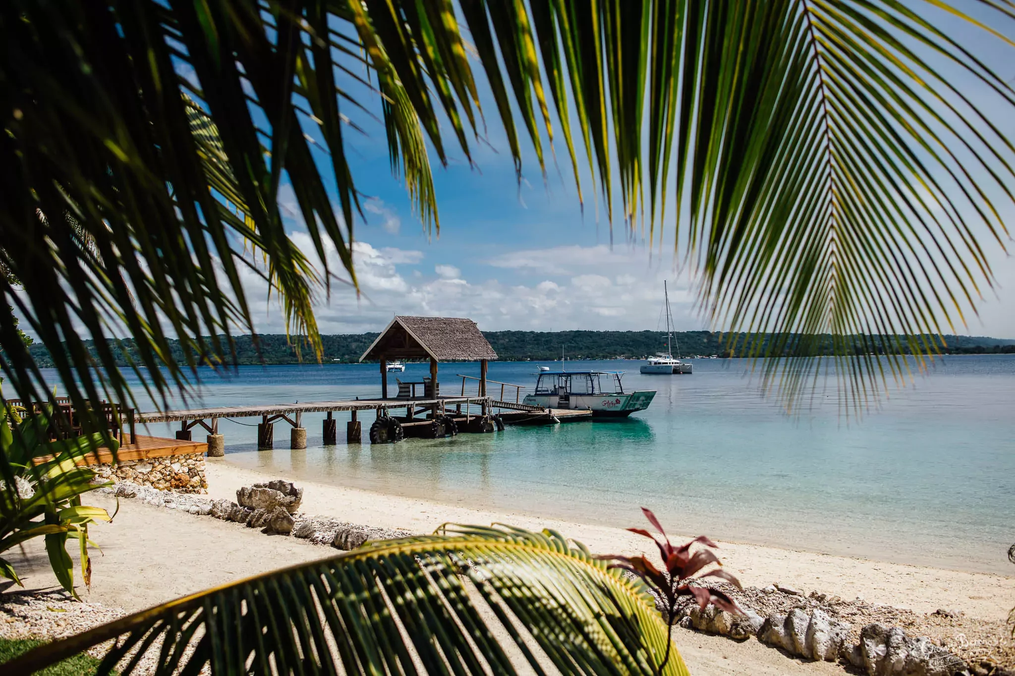 A tropical beach scene with sandy shore, green palm leaves in the foreground, a small wooden dock with a thatched roof over the water, and sailboats anchored in the calm blue water. Hills are visible in the background under a partly cloudy sky.