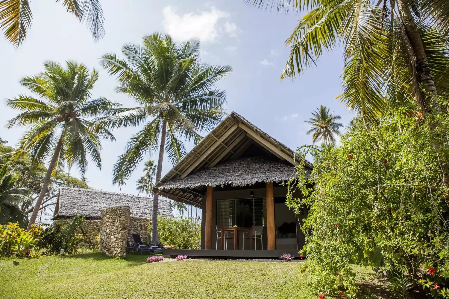 A tropical hut with a thatched roof on a raised wooden deck, surrounded by lush greenery and tall palm trees under a partly cloudy sky.