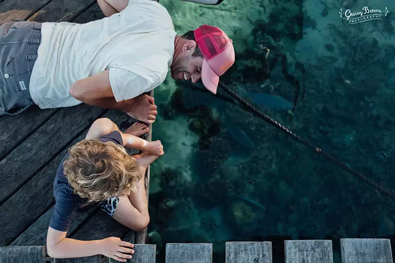 A man and a young girl are sitting on a wooden dock by the water. The man is wearing a white shirt and a red cap, leaning over and smiling as he points a stick into the clear water. The girl has curly blonde hair, is wearing a blue shirt, and is watc