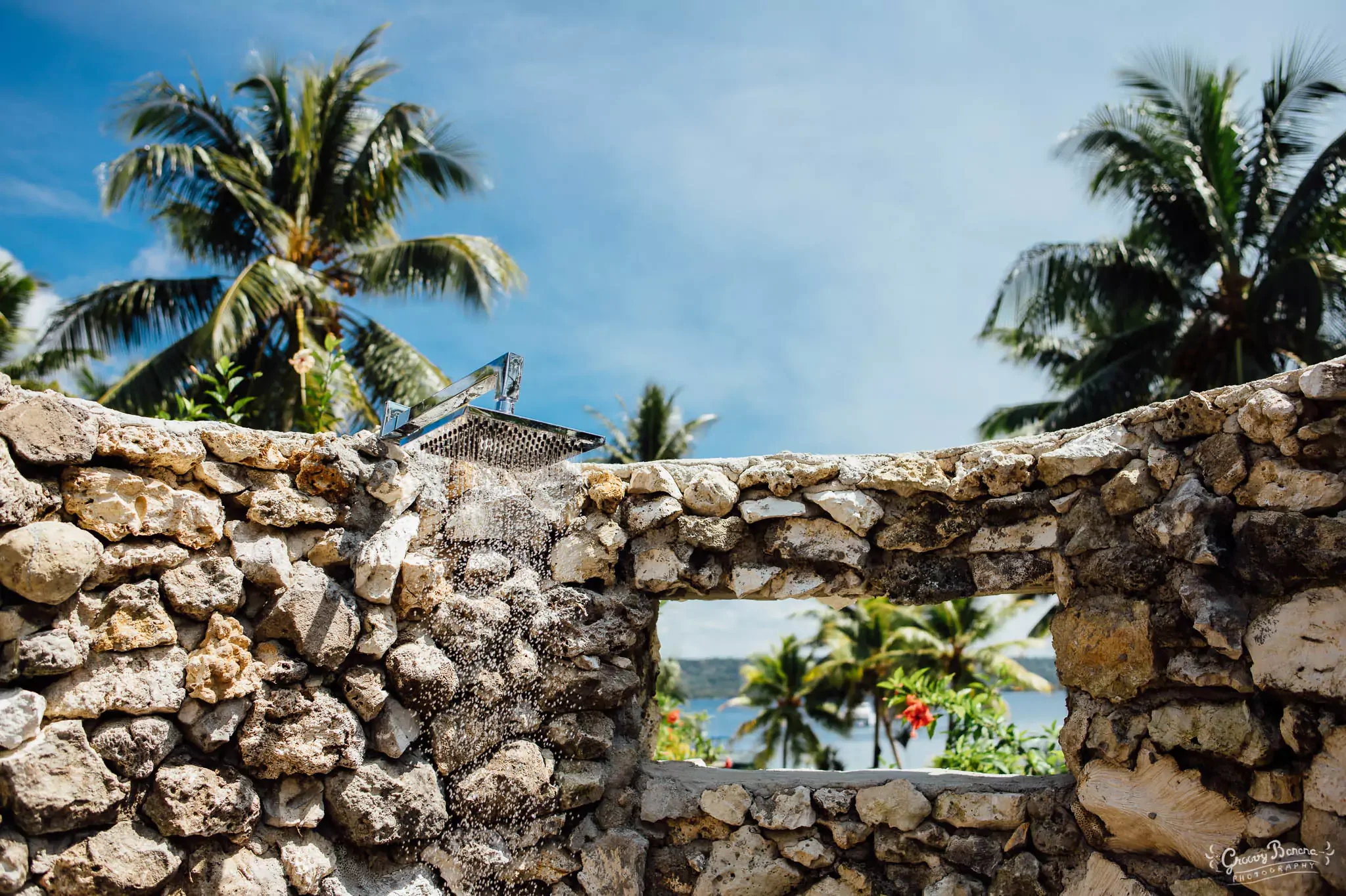 A stone wall with a shower head mounted on top, overlooking palm trees, flowers, and a blue sky.