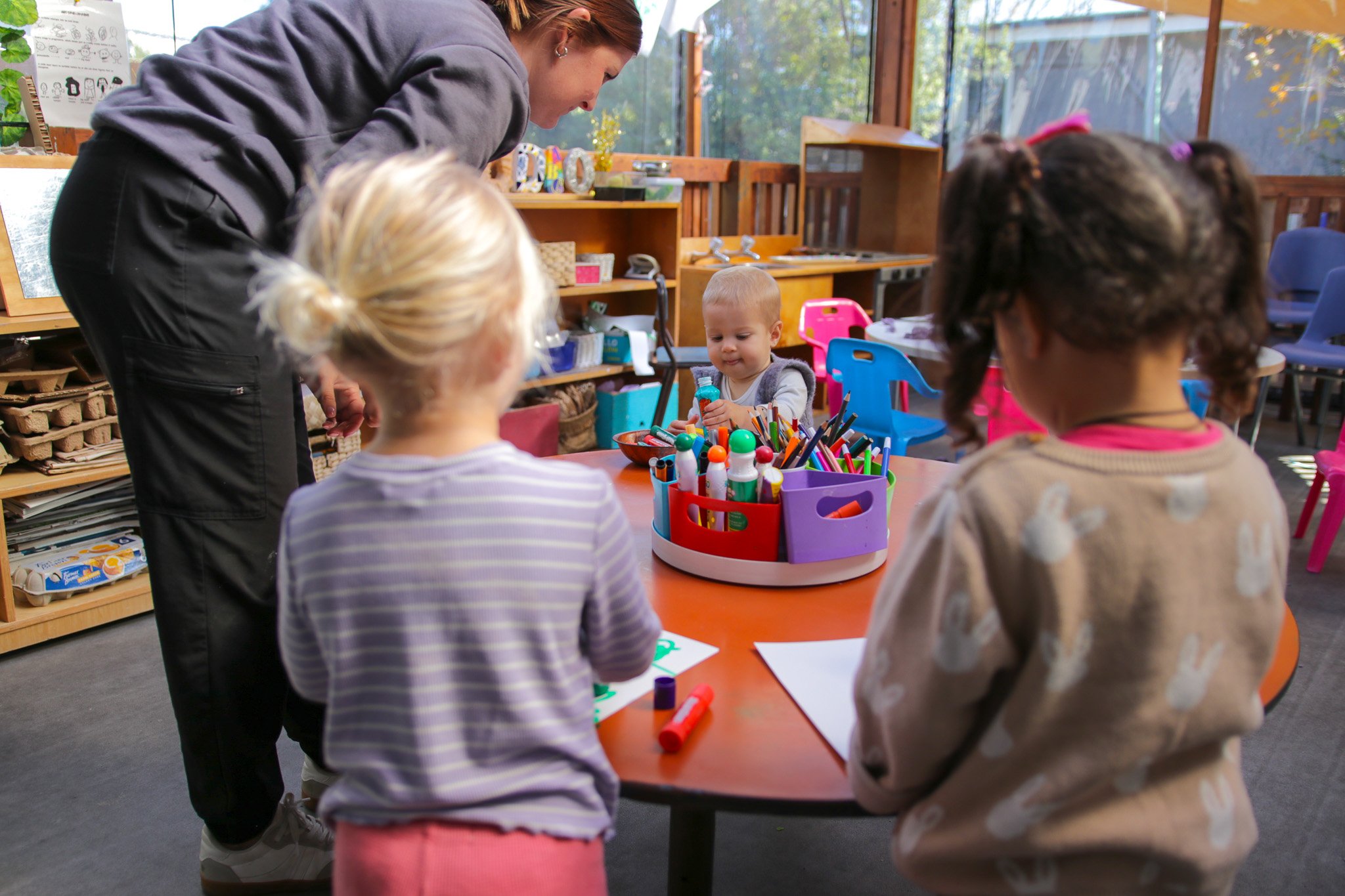 A teacher and four children around a table with art supplies in a classroom, with shelves, colorful chairs, and large windows in the background.