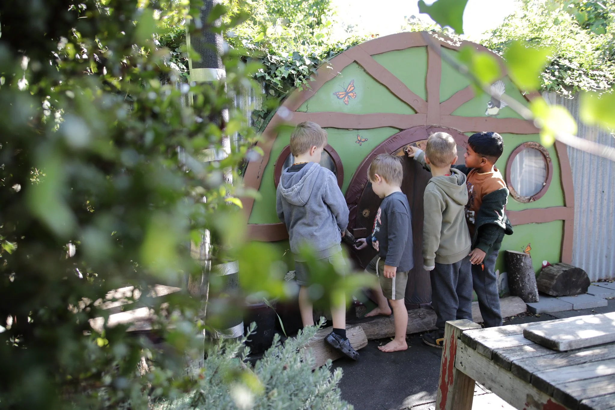 Four children standing in line, observing or playing with a small wooden house with butterfly and bird decorations, surrounded by greenery and outdoor wooden furniture.