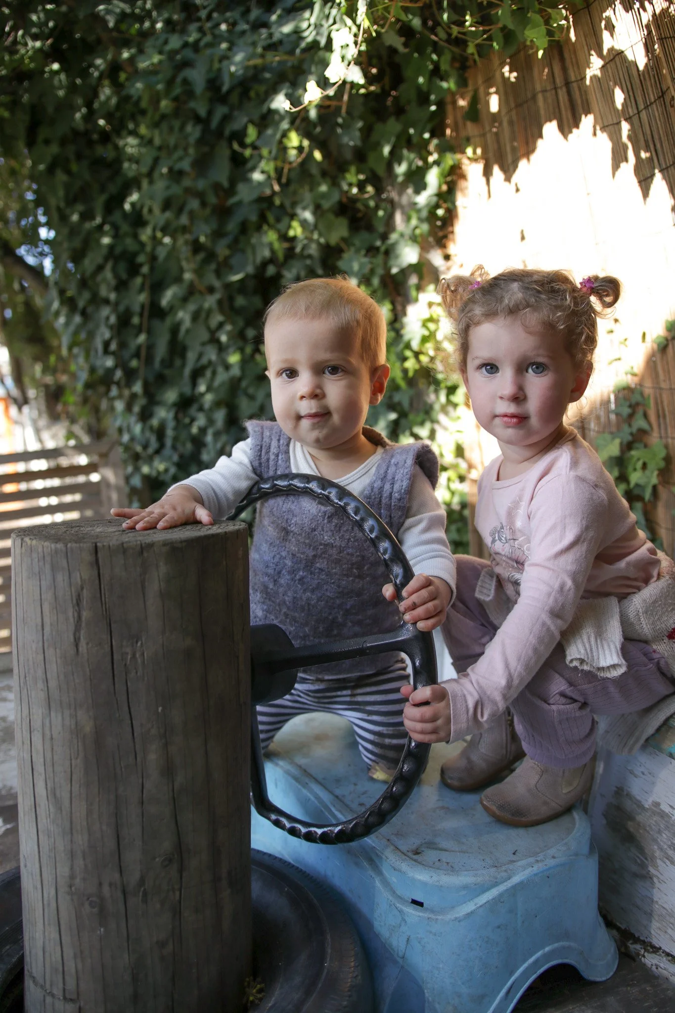 Two young children, a boy and a girl, playing with a large snake at an outdoor setting, surrounded by plants and wooden fencing.