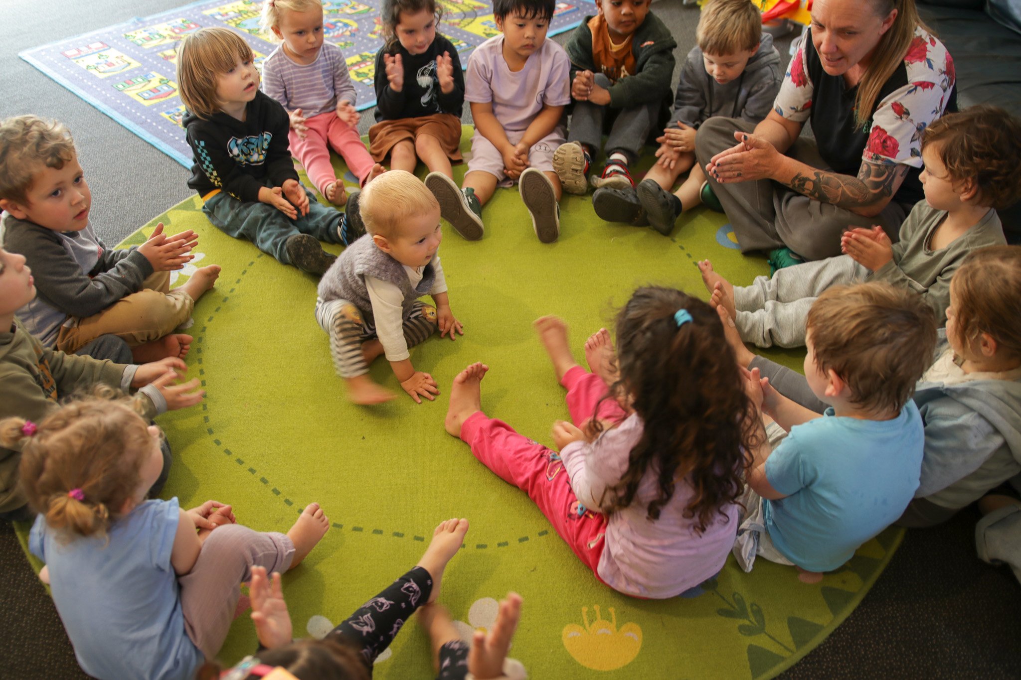 A group of young children sitting on a green circular rug, listening to a woman with long blonde hair and tattoos who is talking to them in a classroom. There is a colorful play mat in the background.