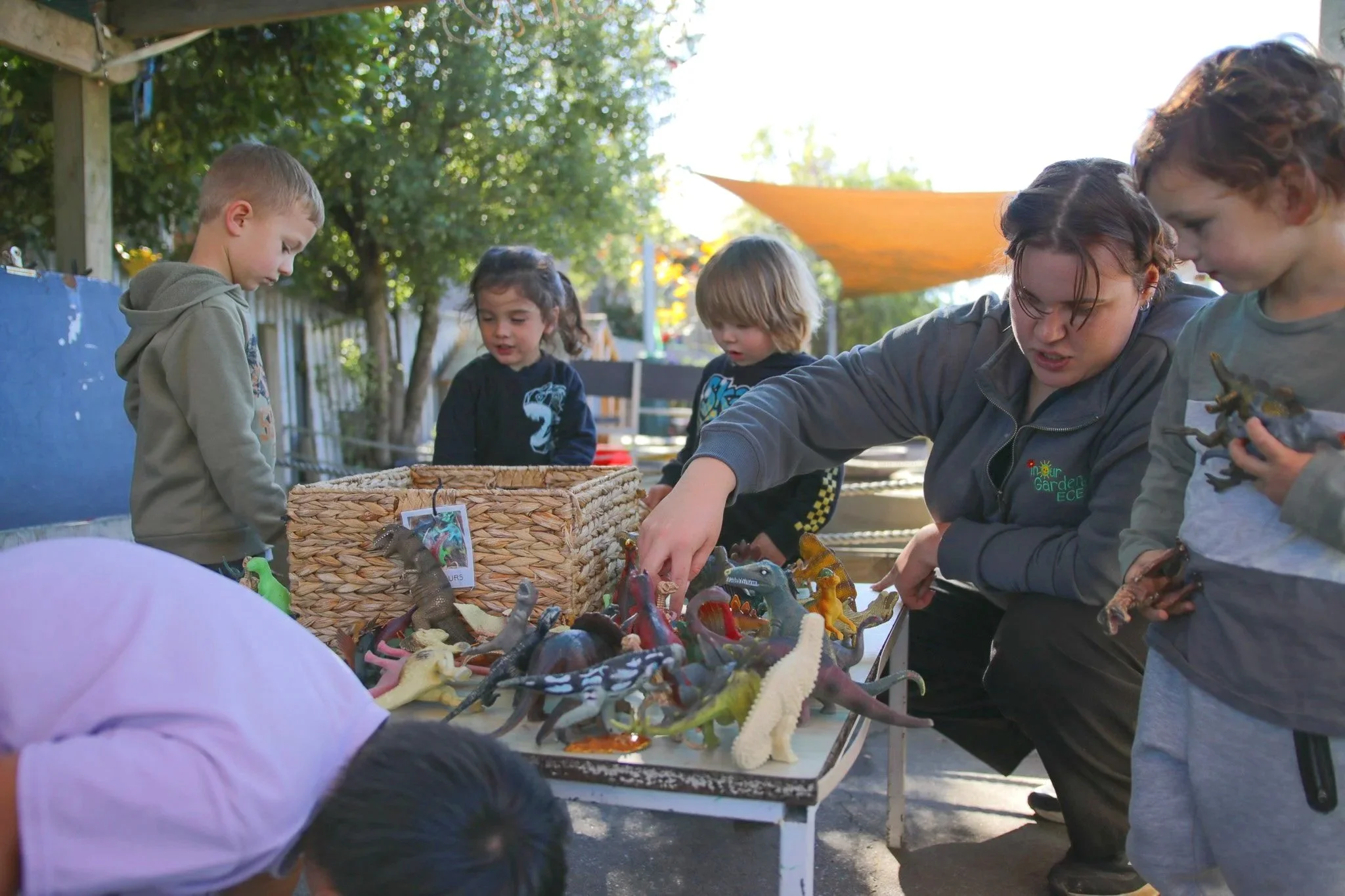 Children and an adult looking at dinosaur figurines displayed on a table outdoors.