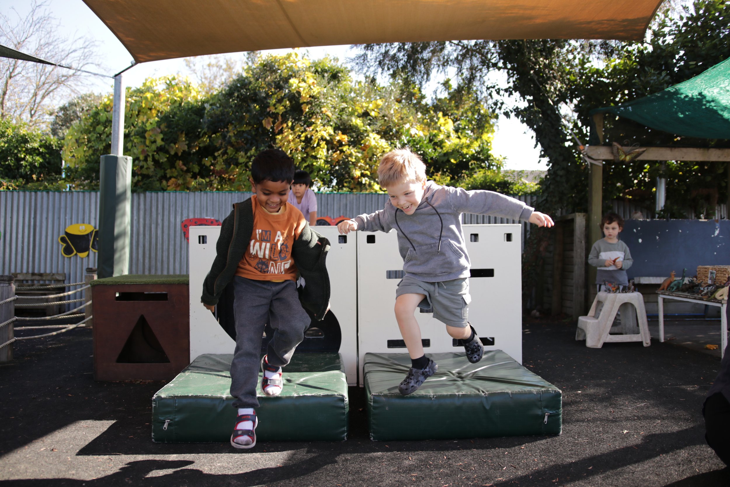Two young boys are jumping off a small platform at an outdoor playground, smiling and having fun, with other children in the background and surrounded by trees and play equipment.