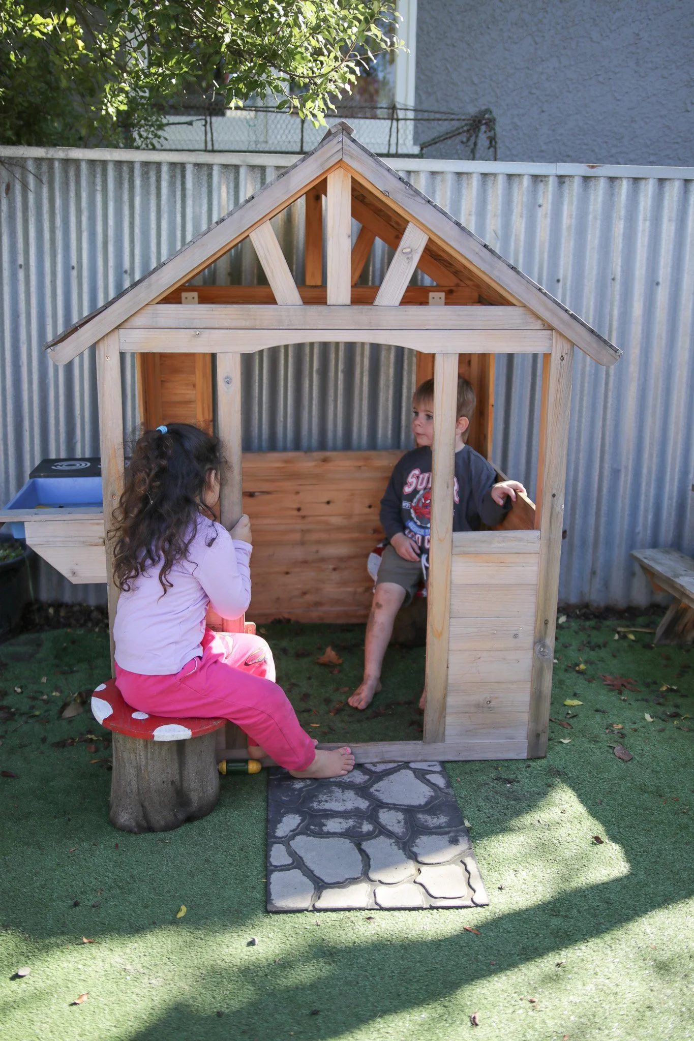 Two young children, a girl and a boy, playing in a small wooden playhouse in a backyard. The girl, wearing pink pants and a light purple shirt, is sitting on a small stool with a red top, and the boy, wearing a gray shirt with a red graphic and shorts, is standing inside the playhouse near the doorway.