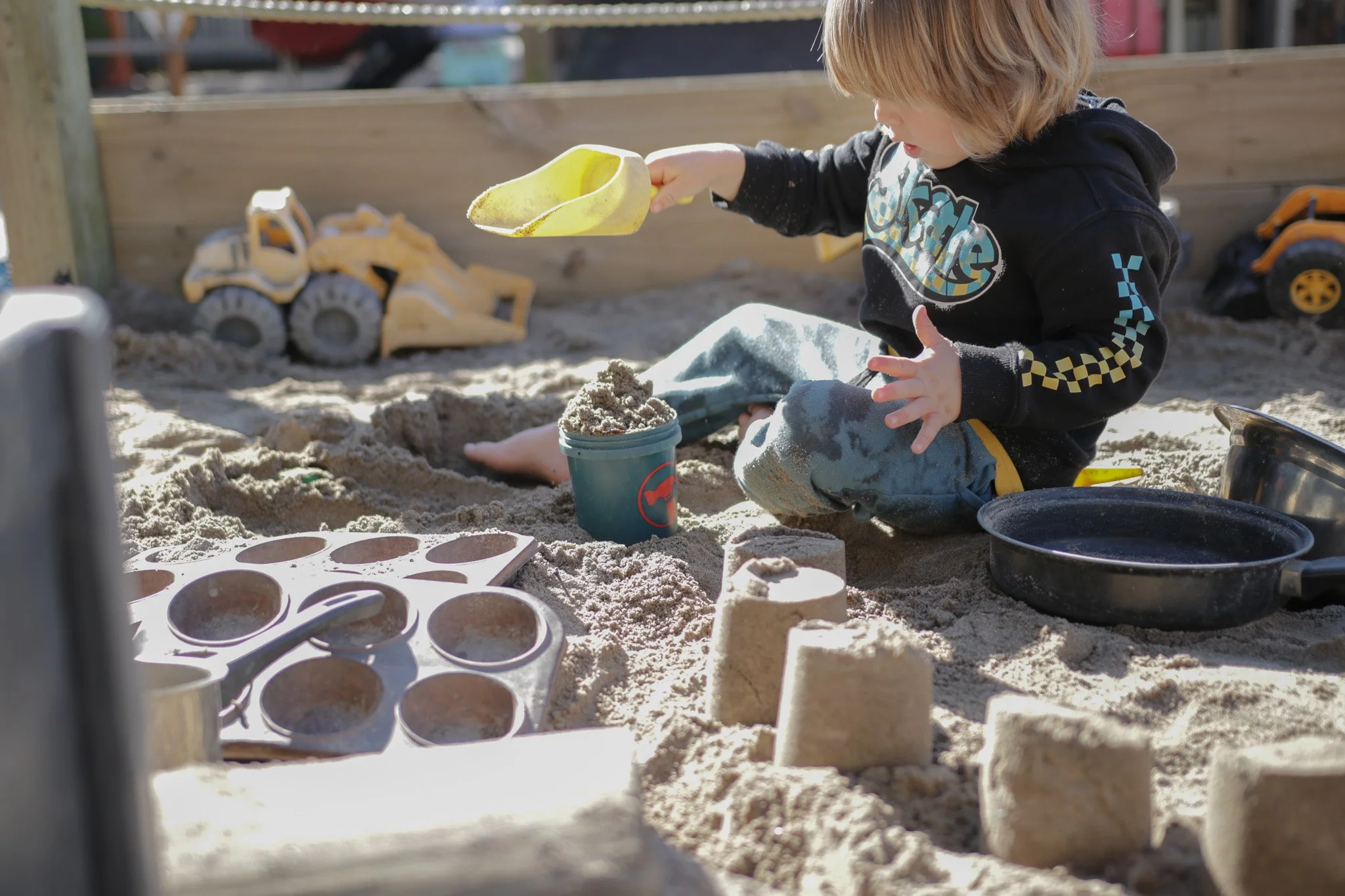 A young boy playing in a sandbox, filling a yellow mold with sand. There are toy construction vehicles, a black container, and a muffin tray with circular molds in the sand. A wooden fence is in the background.