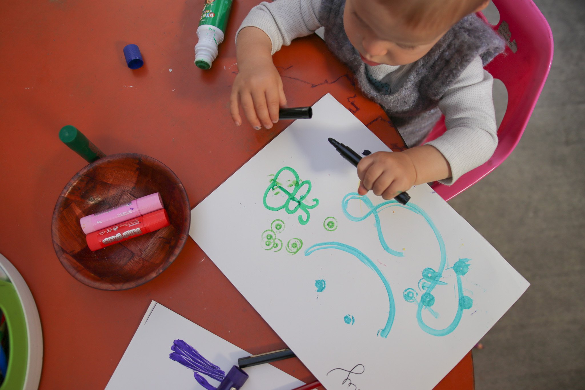A young child sitting at a table drawing with colorful markers on a piece of white paper, with various markers and drawing supplies around them.