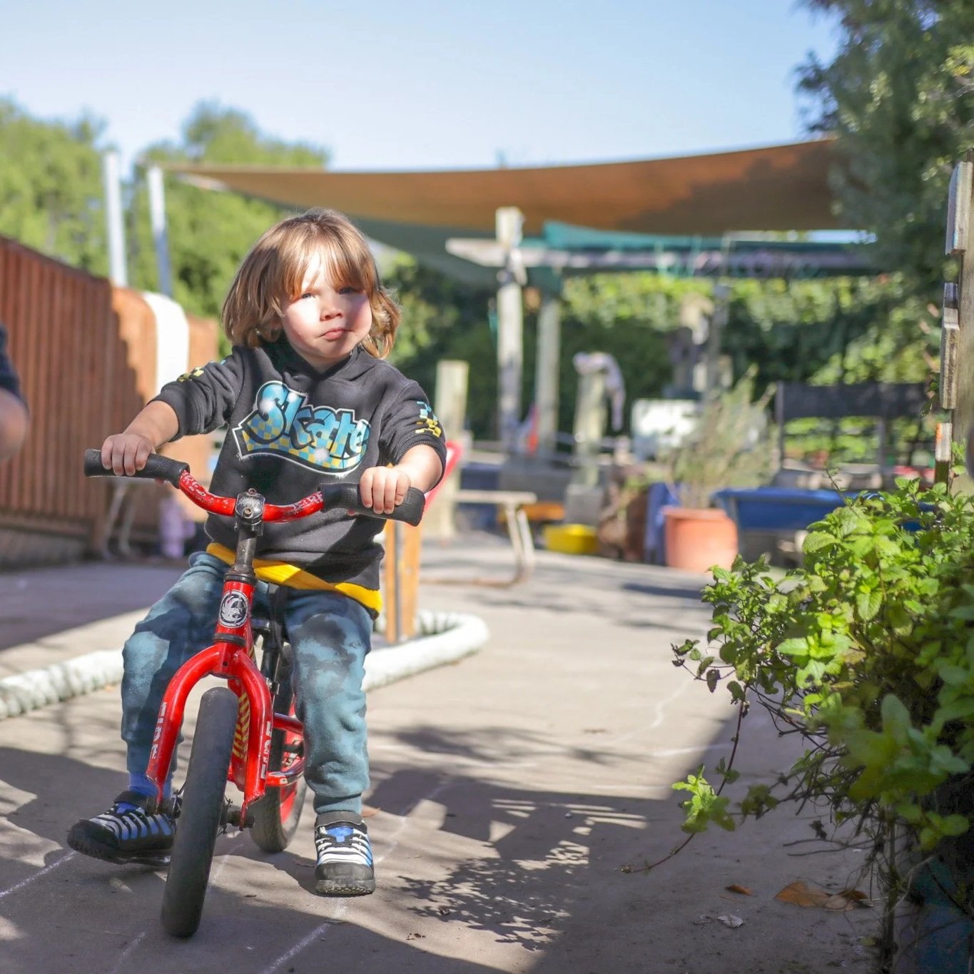 A young child riding a small red bicycle outdoors on a paved path with greenery and outdoor furniture in the background on a sunny day.