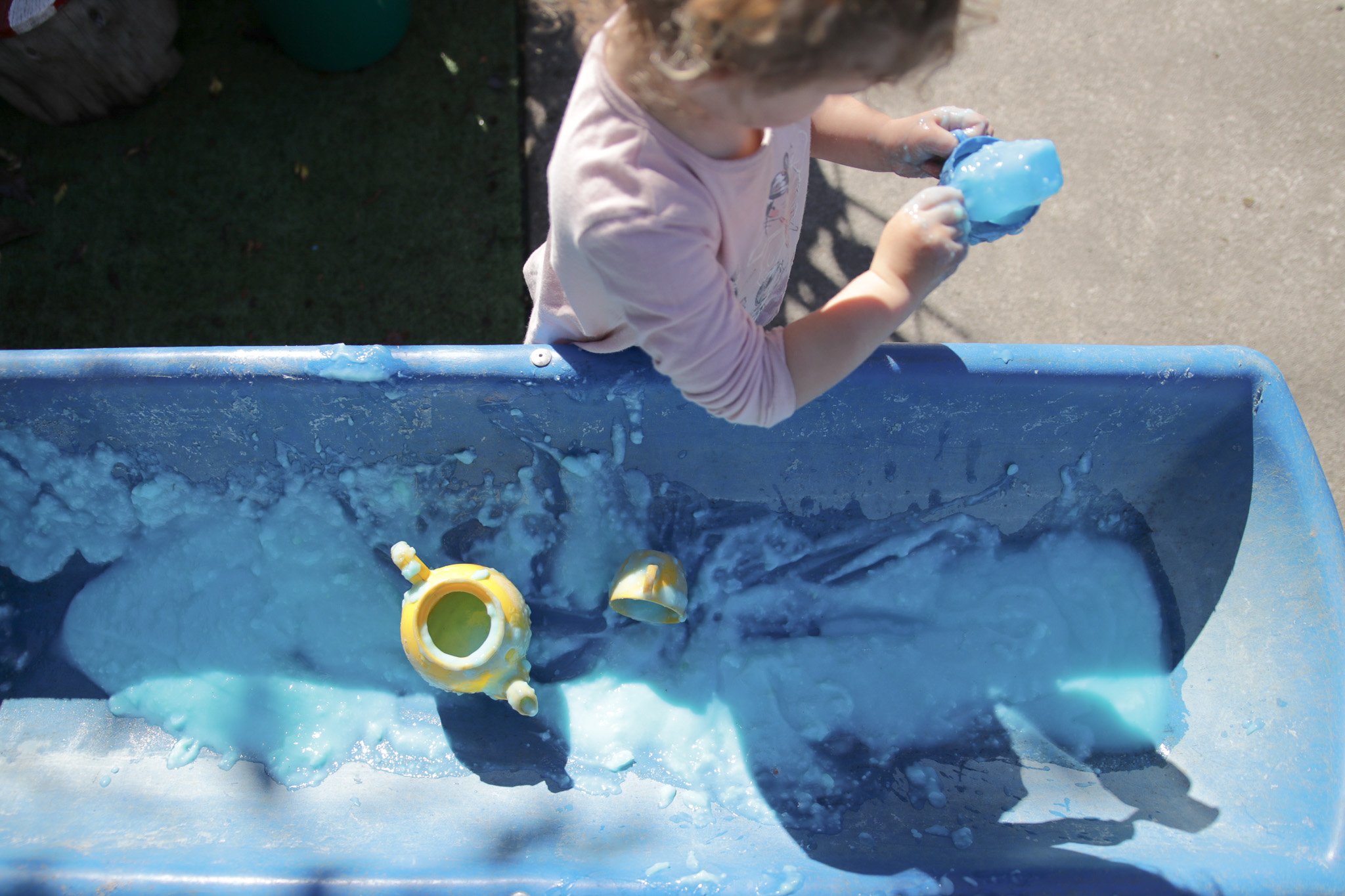 A young child playing with blue slime at an outdoor table, with slime mess on the table surface and yellow slime containers nearby.