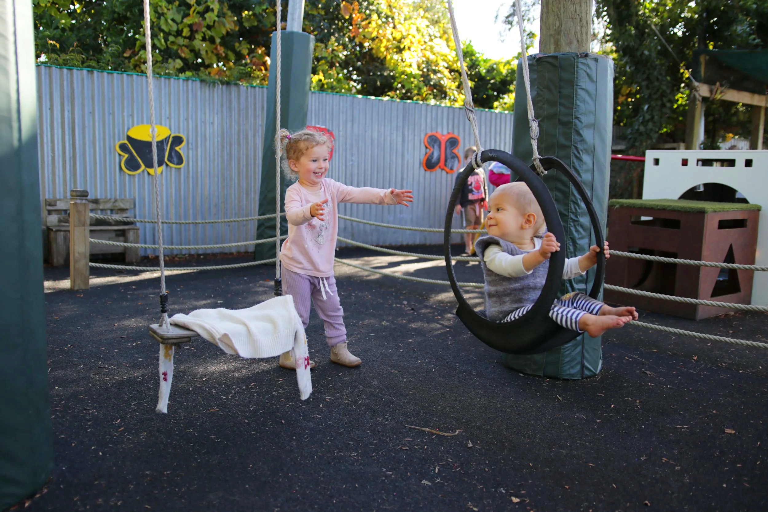 Two young children playing outdoors in a playground enclosed by a metal fence decorated with colorful butterfly and bee decorations. One girl is standing, smiling, and reaching towards a baby sitting in a black infant swing, which is suspended from a swing set. The baby looks up at the girl with a content expression. The background shows trees, wooden structures, and other children.