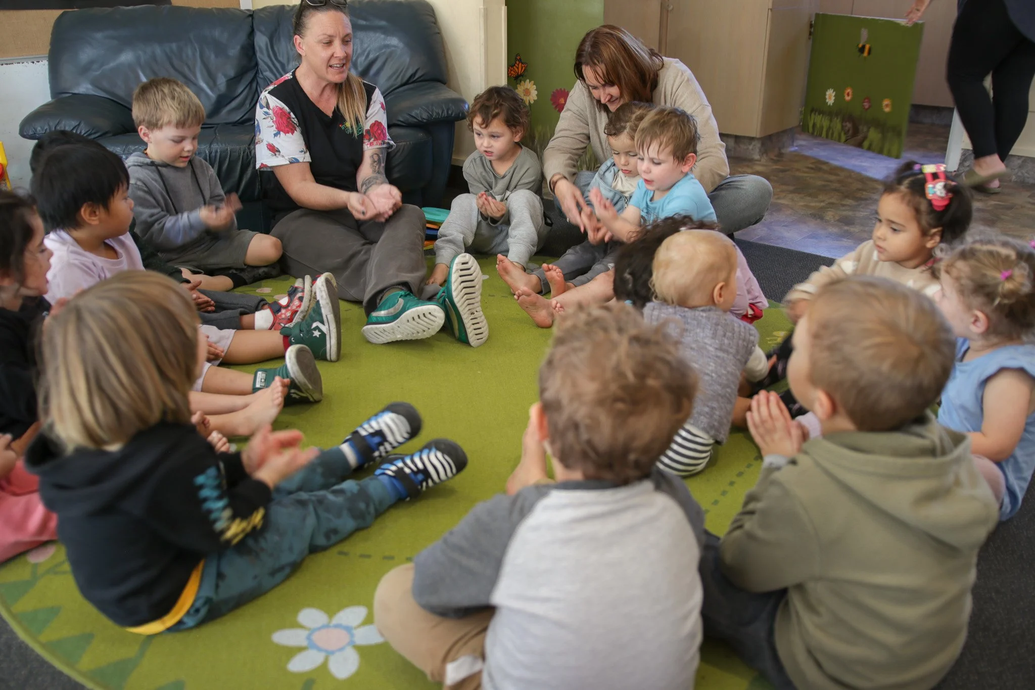 A group of young children and two adult women sitting on a green carpet with colorful flower patterns in a circle, engaging in a conversation or activity. The children are listening attentively and clapping, while the women are smiling and talking to them.