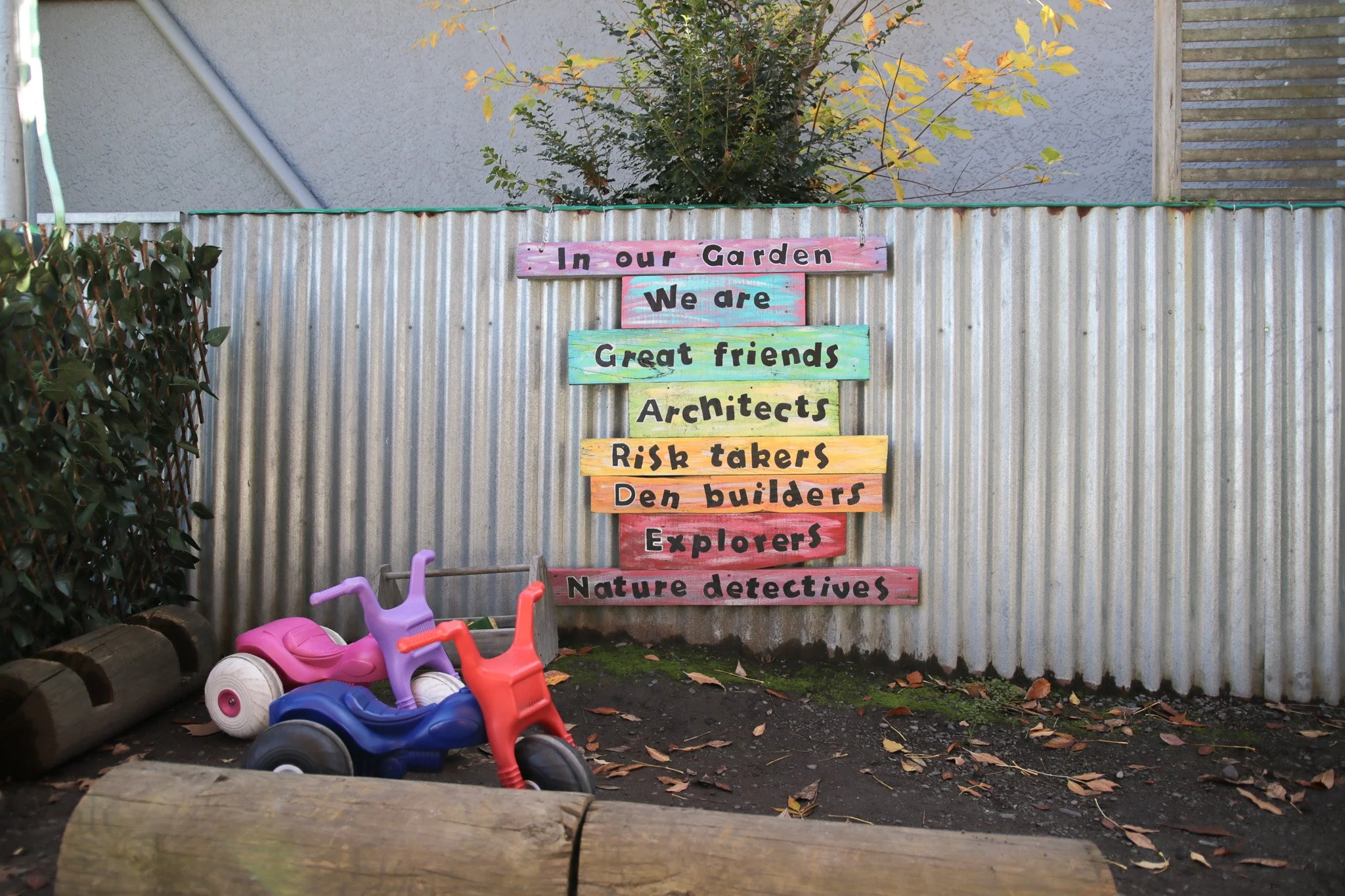 Colorful wooden sign with phrases in black lettering, including 'In our Garden,' 'We are,' 'Great friends,' 'Architects,' 'Risk takers,' 'Den builders,' 'Explorers,' 'Nature detectives,' mounted on a corrugated metal fence. In front, children's toy cars are placed on dark soil with fallen leaves, with a hedge on the left and a tree with yellowing leaves behind.