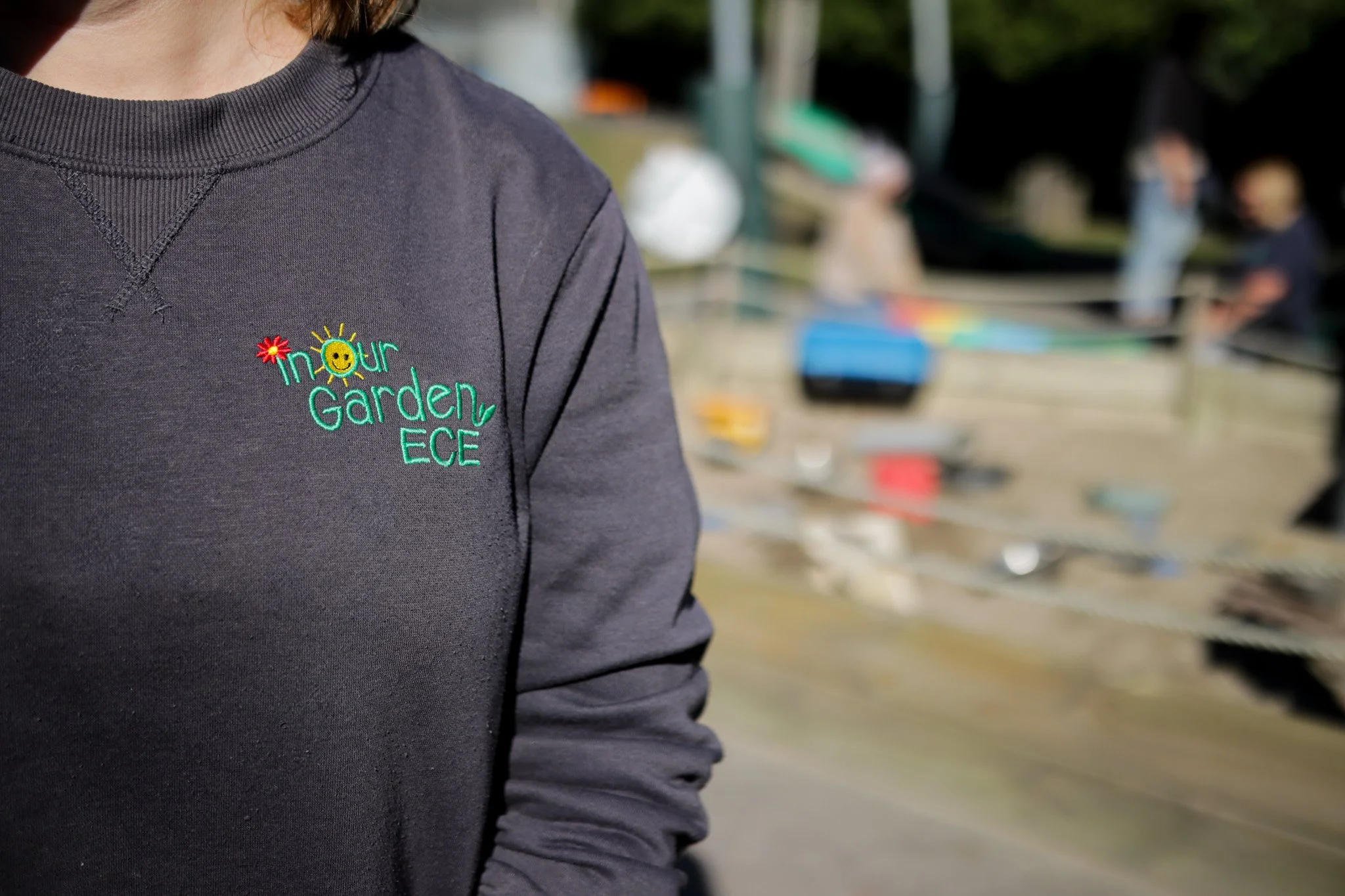 Close-up of a person wearing a black sweatshirt with colorful embroidery that says "In Your Garden EC" and has a smiling sun with sun rays and a red flower. The background shows people and outdoor objects, slightly blurred.