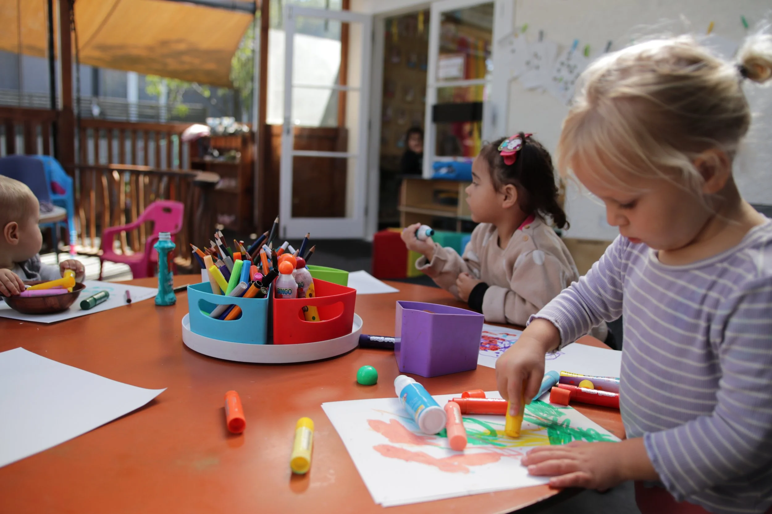 Children creating artwork at a table with markers, crayons, and paper in a classroom or creative space.