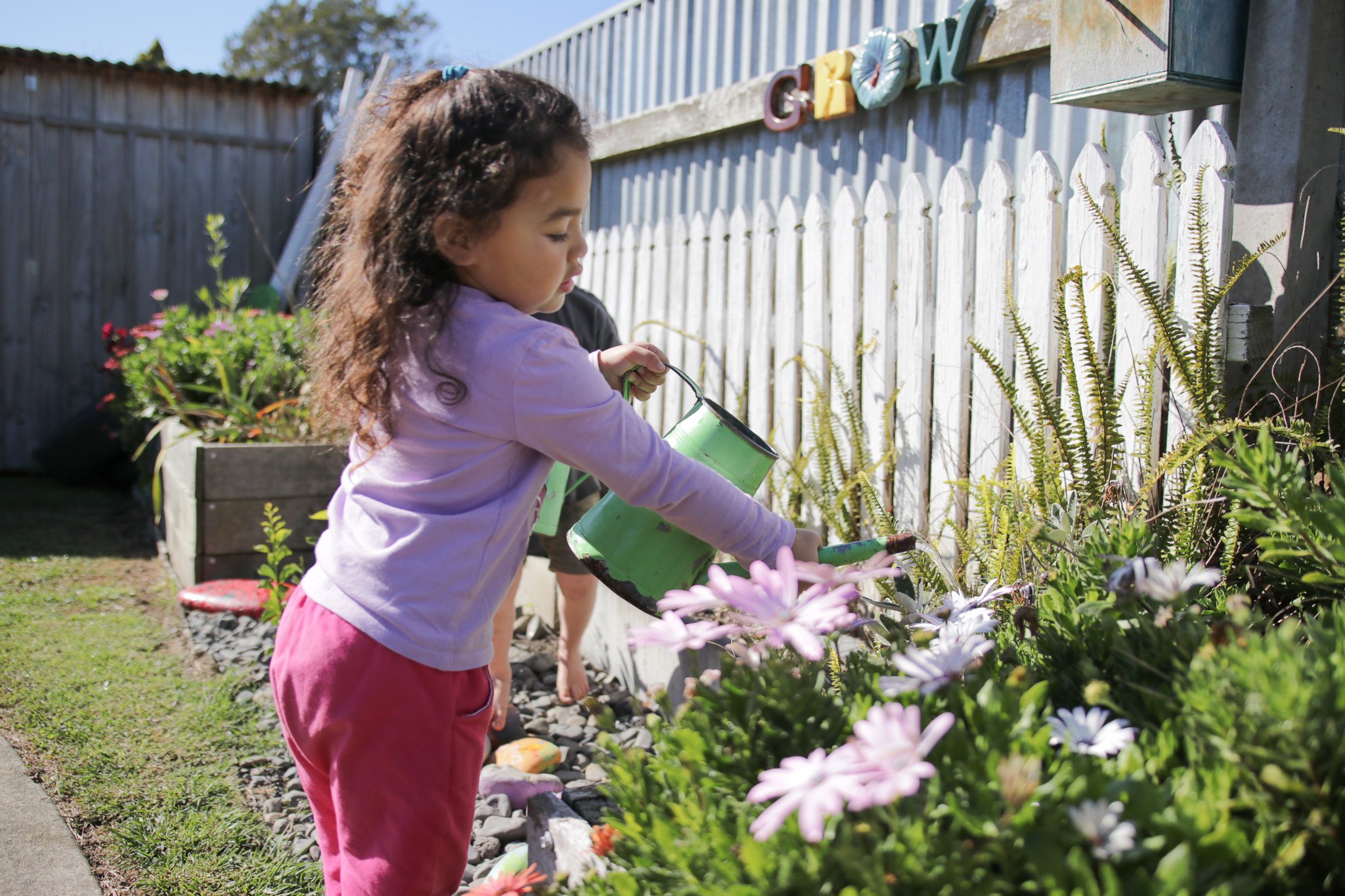 Young girl watering flowers in a garden next to a white picket fence with colorful decorative letters on top.