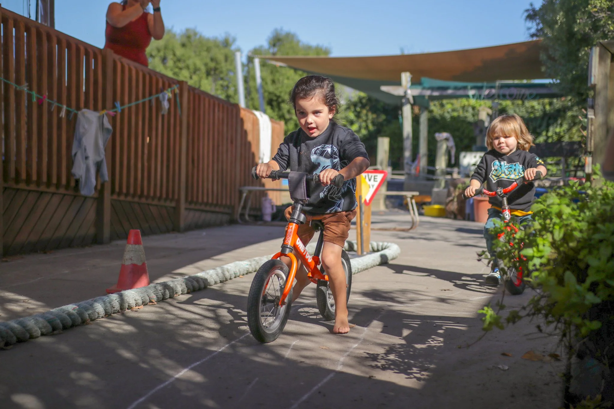 Young boy riding a bicycle barefoot on a sunny backyard pathway, with another child on a bike behind him, and a woman standing on a wooden deck watching. The area is decorated with colorful clothespins and laundry hanging, with trees and outdoor furniture in the background.