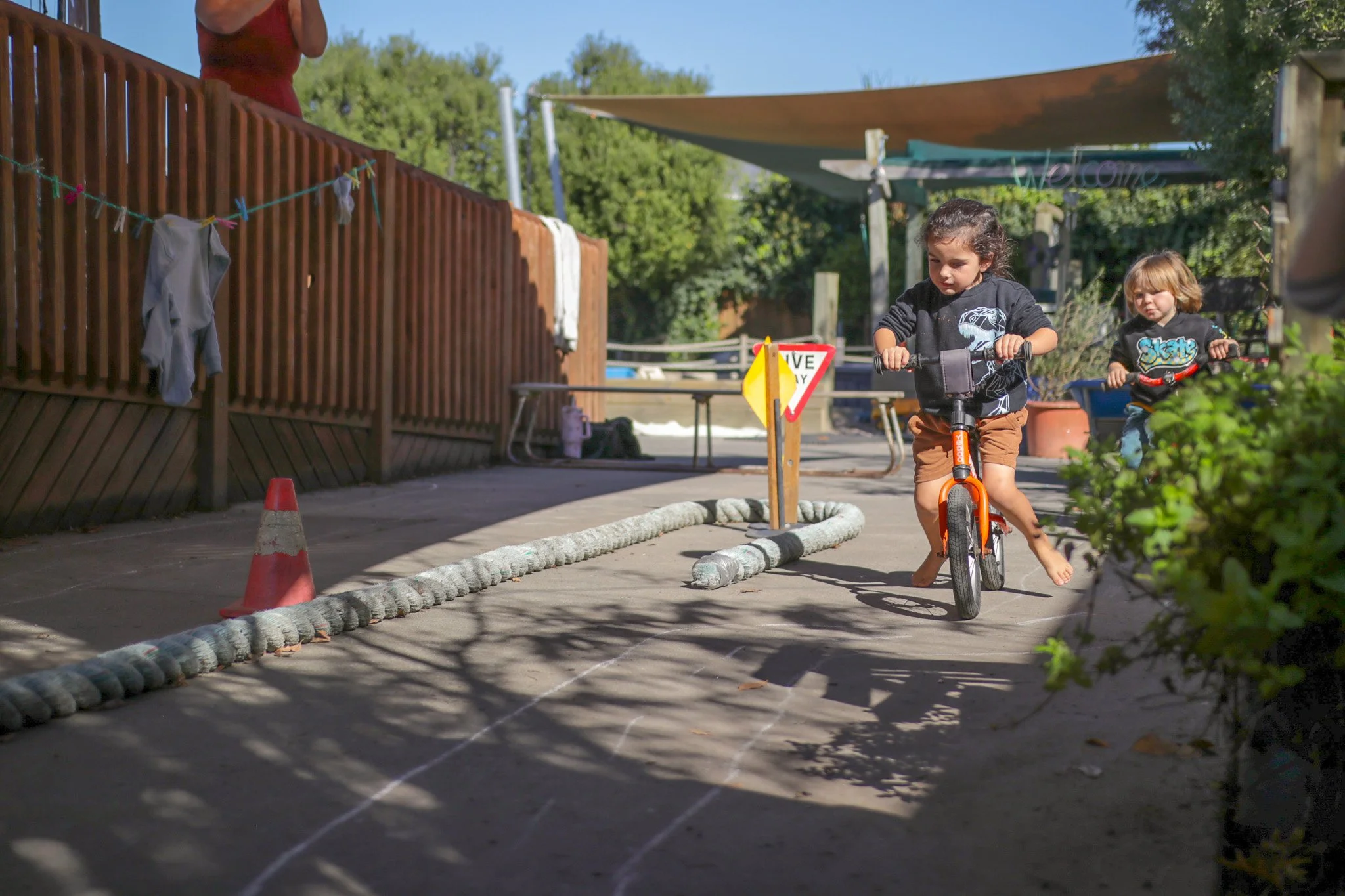 Two young children, a girl and a boy, riding orange bicycles in a backyard, with a wooden fence, trees, and a shaded patio in the background.