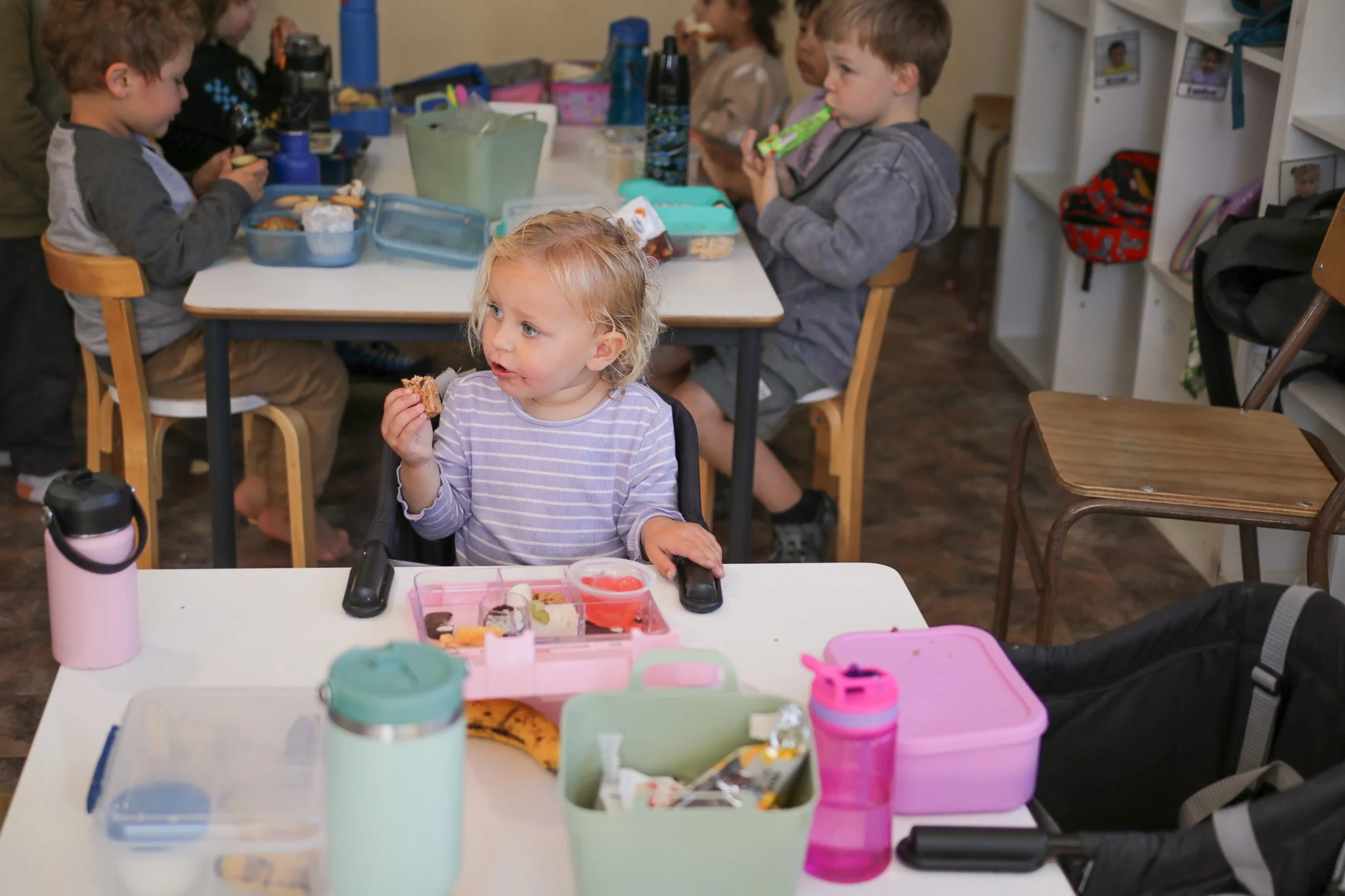 Young children eating lunch at a school cafeteria table with lunchboxes and water bottles in front of them.